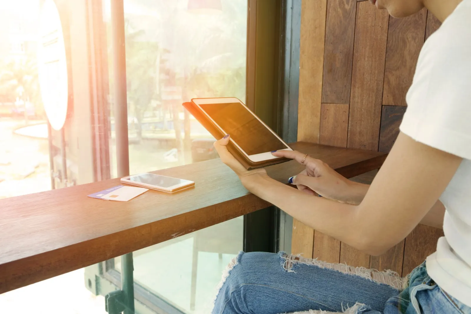 A woman uses a tablet by a sunlit cafe window, with a smartphone on the table.